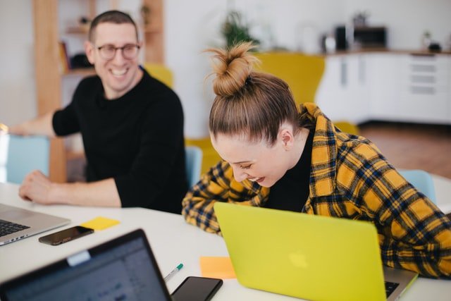 Man and a woman sitting in a meeting room and laughing with their laptops in front of them